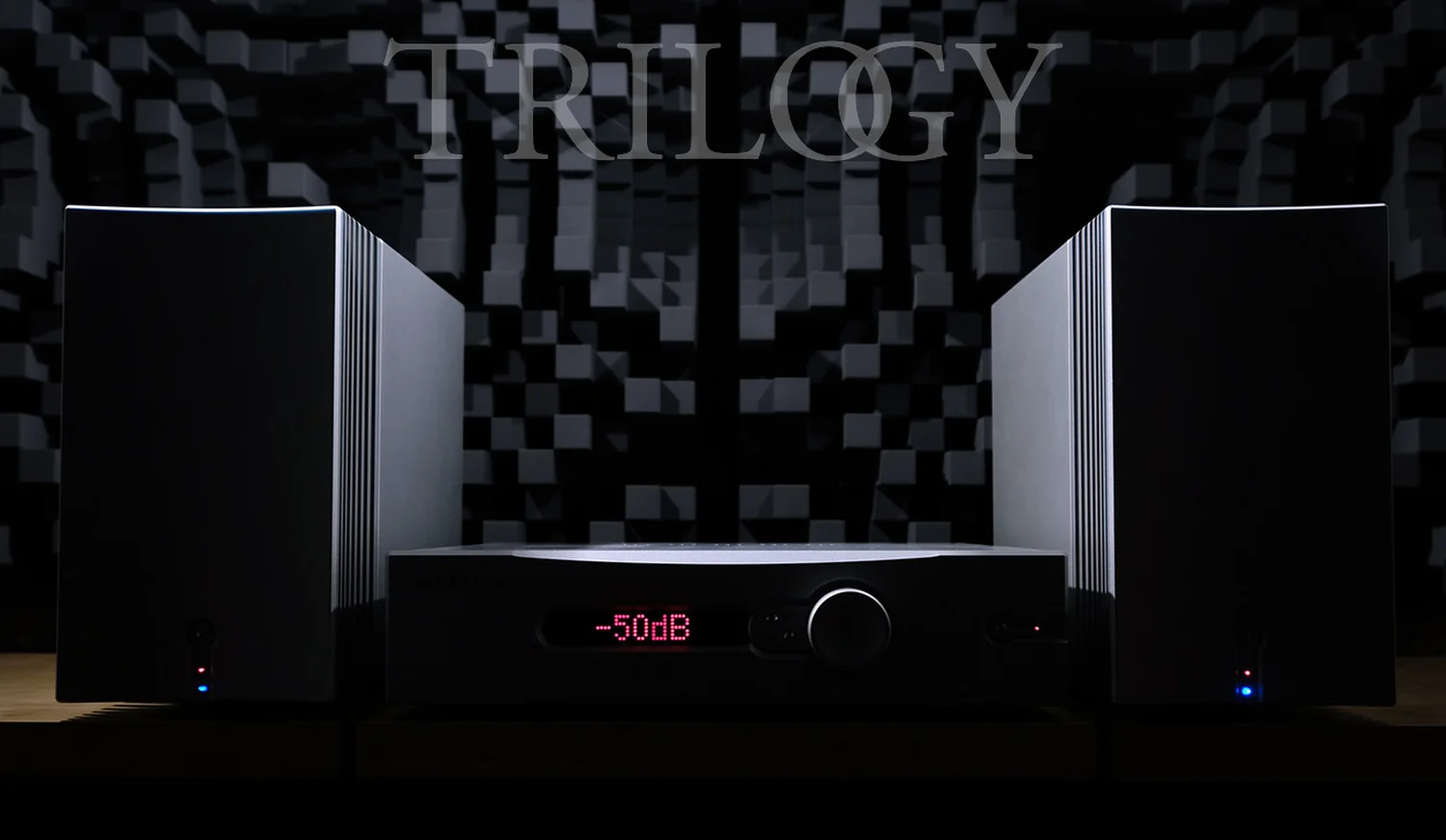 Un ensemble Trilogy Audio avec un préamplificateur 995R au centre central affichant « -50db » flanqué de deux amplificateurs mono 995R sur fond de diffuseurs gris (par 1877.audio)