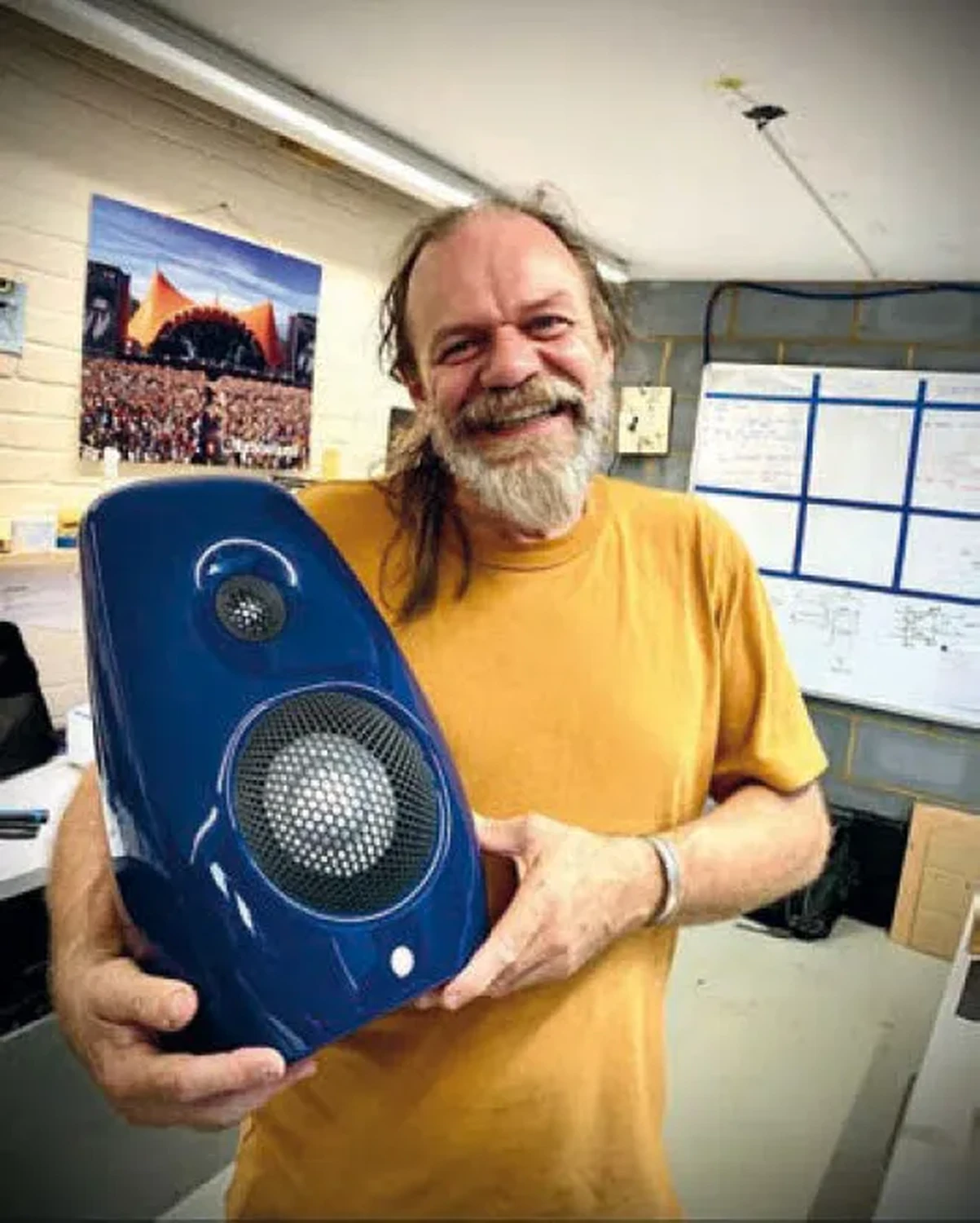 Laurence Dickie, un homme barbu souriant en T-shirt jaune, tient une enceinte Vivid Audio Kaya S12 bleue électrique dans une pièce avec une photo de festival de musique en arrière-plan (par 1877.audio)