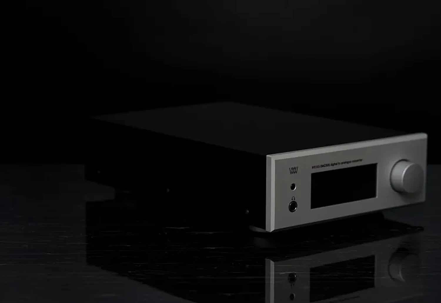 Un convertisseur audio numérique Weiss DAC501 avec un panneau avant de couleur aluminium argenté et côté métalliques noirs placé sur une surface noire brillante et sur fond sombre (par 1877.audio)