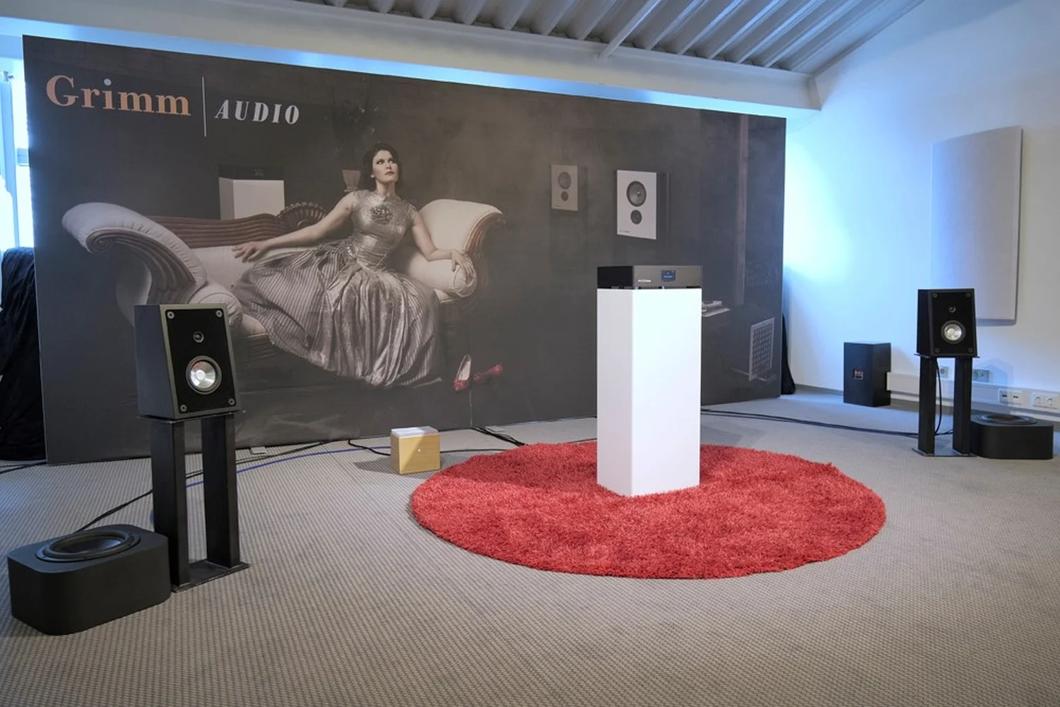 Une pièce de démo sur le salon de Munich 2023 avec le DAC Grimm Audio MU2 sur une colonne blanche brillante, une paire enceintes basique avec leur caisson avec en fond un grand décor présentant une femme assise sur un canapé (par 1877.audio)