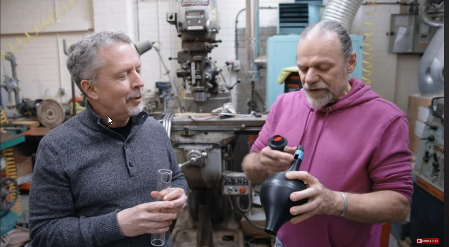 Deux hommes engageant une conversation dans un atelier ; celui de droite est un journaliste de SoundStage qui tient un tube transparent et celui de droite est Laurence Dickie qui tient lui un "absorber" de son invention, qu'il utilise dans les enceintes Vivid Audio (par 1877.audio)