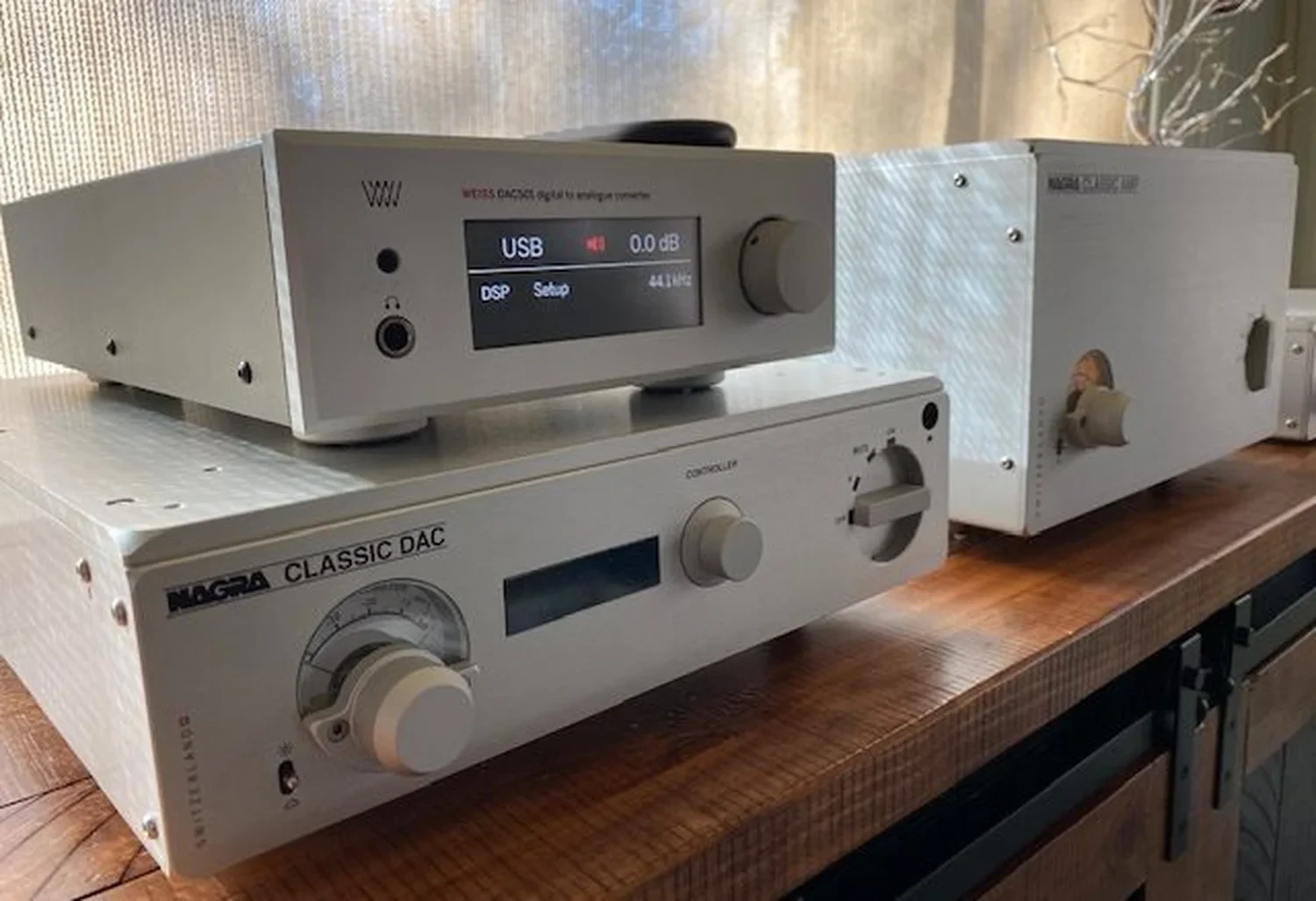 Deux composants audio haut de gamme, dont un Weiss DAC501 posé sur un préamplificateur Nagra et à coté d'un amplificateur de puissance Nagra également, le tout sur un meuble en bois exotique (par 1877.audio)